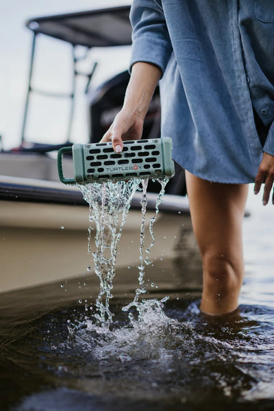 Person holding a waterproof speaker in water near a boat